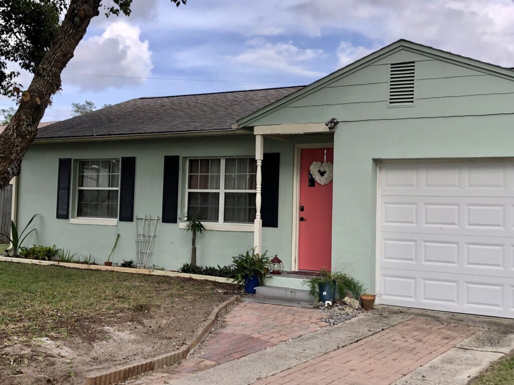 1950s Cottage, Front of home, pink door, black shutters, mint green painted stucco, white single car garage
